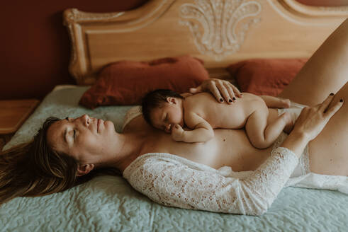 Side view of adorable newborn lying on naked torso of calm mother with closed eyes in bedroom - ADSF32636