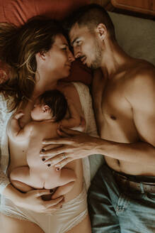 From above naked man embracing happy wife with newborn while lying on bed after breastfeeding - ADSF32637