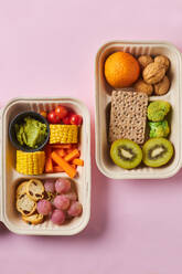 From above of lunch boxes with healthy food including crackers carrot sticks grapes cherry tomatoes with kiwi broccoli walnut and tangerine on pink background - ADSF32663