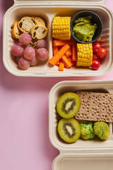 Von oben von Lunch-Boxen mit gesunden Lebensmitteln einschließlich Cracker Karottensticks Trauben Kirschtomaten mit Kiwi Brokkoli Walnuss und Mandarine auf rosa Hintergrund - ADSF32664