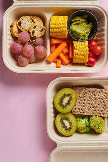 From above of lunch boxes with healthy food including crackers carrot sticks grapes cherry tomatoes with kiwi broccoli walnut and tangerine on pink background - ADSF32664