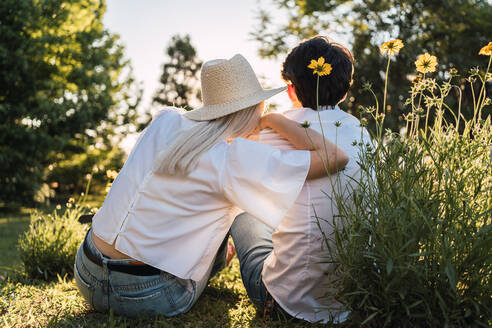 Rückenansicht eines nicht erkennbaren liebenden Mannes und einer Frau, die während eines romantischen Dates auf einer Wiese im Wald zusammensitzen - ADSF32666