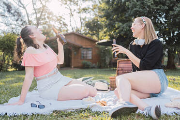 Full body of cheerful young female friends in casual clothes laughing while drinking beer sitting on blanket during picnic in nature on sunny summer day - ADSF32670