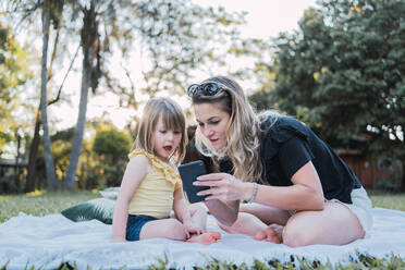 Full body of young barefooted female with long blond hair in casual clothes sitting on blanket and showing smartphone to curious little daughter during holidays in countryside - ADSF32672