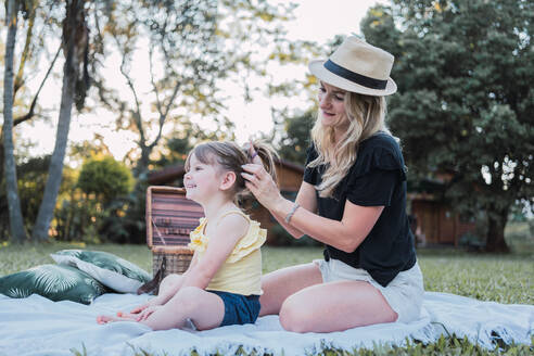 Full body of happy young mother with long blond hair in casual clothes and straw hat making hair of adorable positive little daughter sitting on blanket during picnic in countryside - ADSF32673