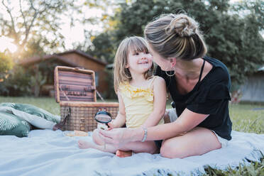 Positive young mother in casual clothes and straw hat hugging cute little daughter and looking together at insect with magnifying glass during picnic in nature - ADSF32675