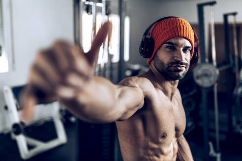 Muscular shirtless Hispanic male athlete in hat and wireless headphones showing shaka gesture during training in gym with various fitness equipment - ADSF32679