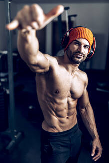 Muscular shirtless Hispanic male athlete in hat and wireless headphones showing shaka gesture during training in gym with various fitness equipment - ADSF32680