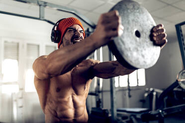Wütender, muskulöser, hemdloser, hispanischer Mann mit drahtlosen Kopfhörern, der laut schreit, während er beim Training im Fitnessstudio eine schwere Scheibe hebt - ADSF32681