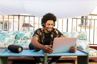 Glücklicher afroamerikanischer Mann, der mit Kopfhörern Musik hört und auf seinem Netbook surft, während er auf einer Terrasse in der Stadt auf der Couch sitzt - ADSF32685