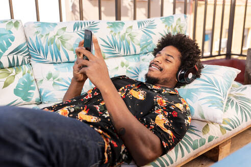 Seitenansicht eines männlichen Afroamerikaners, der auf einer Terrasse liegend Musik hört und Textnachrichten auf seinem Mobiltelefon verschickt - ADSF32688