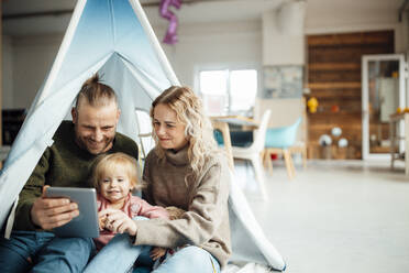 Happy family sitting inside tent using digital tablet at home - GUSF06655