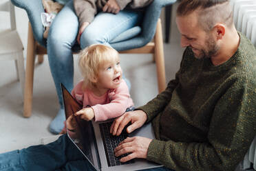 Daughter pointing at laptop on father's lap with mother in background - GUSF06666