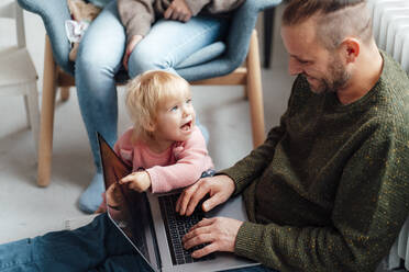 Tochter zeigt auf Laptop auf dem Schoß des Vaters mit Mutter im Hintergrund - GUSF06666