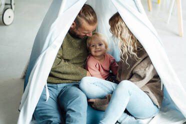 Playful daughter sitting with parents inside tent at home - GUSF06678