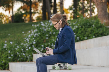 Businessman with laptop using mobile phone on skateboard - JRVF02240