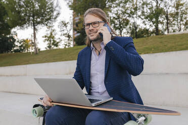 Businessman with skateboard and laptop talking on mobile phone at steps - JRVF02246