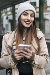 Smiling woman with smart phone leaning on glass wall - JRVF02267