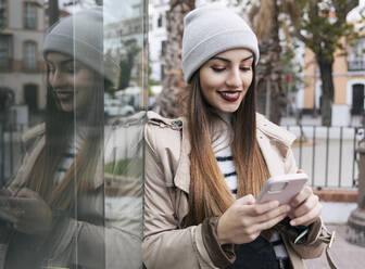 Woman using smart phone leaning on glass wall - JRVF02269