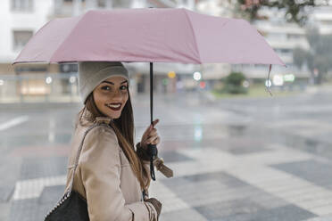 Woman looking over shoulder holding umbrella at city street - JRVF02274