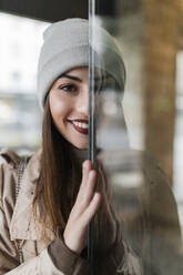 Young woman in knit hat peeking behind glass wall - JRVF02277