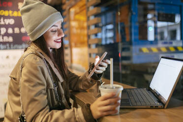 Frau, die ein Smartphone benutzt und ein Getränk am Laptop in einem Café hält - JRVF02292