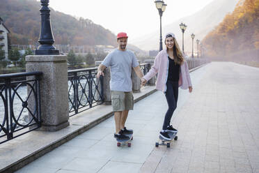 Paar hält sich an den Händen und fährt gemeinsam Skateboard auf dem Fußweg - OMIF00229