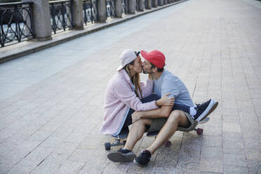 Affectionate couple kissing each other sitting on skateboard at footpath - OMIF00232