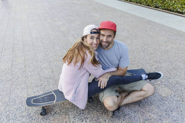 Frau umarmt ihren Freund auf dem Skateboard sitzend auf dem Gehweg - OMIF00235