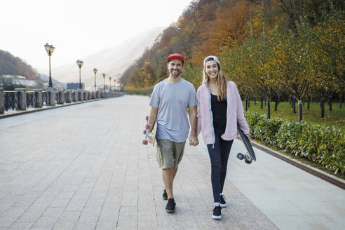 Lächelndes Paar mit Skateboards, das sich an den Händen hält und gemeinsam auf dem Fußweg geht - OMIF00240