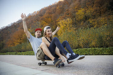Verspieltes Paar, das zusammen auf dem Fußweg Skateboard fährt - OMIF00248