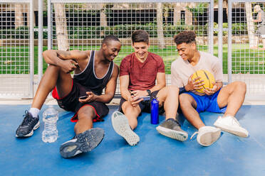 Fröhliche junge männliche Athleten in Sportkleidung, die auf Smartphones surfen und lachen, während sie in der Nähe des Netztores auf dem Sportplatz sitzen - ADSF32690