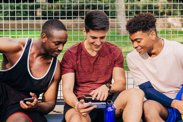 Fröhliche junge männliche Athleten in Sportkleidung, die auf Smartphones surfen und lachen, während sie in der Nähe des Netztores auf dem Sportplatz sitzen - ADSF32691