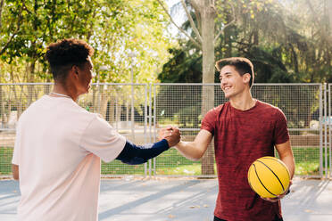 Junge männliche Sportler mit gelbem Basketball schütteln sich die Hände, während sie auf dem Sportplatz im Park stehen - ADSF32693