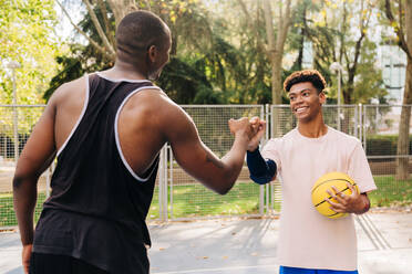 Young ethnic back male athletes with yellow basketball shaking hands while standing on sports ground in park looking at each other - ADSF32694