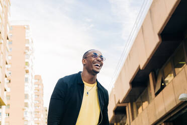 Von unten lächelnder ethnischer Mann mit Brille, der auf dem unscharfen Hintergrund einer Stadtstraße wegschaut - ADSF32700
