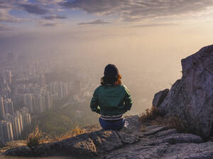 Frau sitzt auf einem Felsen in den Hügeln über Hongkong und blickt auf die Stadt hinunter. - MINF16471
