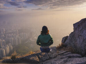 Woman sitting on rock in hills above Hong Kong looking down over the city. - MINF16471