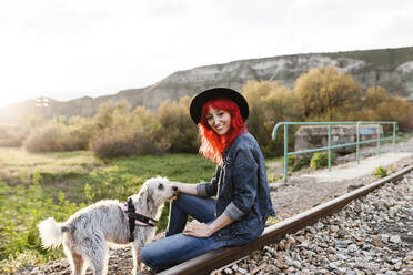 Lächelnde Frau, die einen Hund streichelt, der auf einem Bahngleis sitzt - MRRF01764