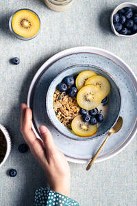 Draufsicht auf eine anonyme weibliche Person beim Frühstück in einer Schüssel mit Müsli, Blaubeeren und Kiwi-Scheiben - ADSF32738