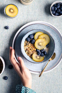 Top view of crop anonymous female with breakfast with bowl with granola and blueberries and kiwi slices - ADSF32738