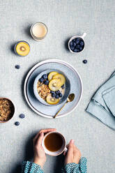 Top view of crop anonymous female warming hand with cup of hot drink at breakfast with bowl with granola and blueberries and kiwi slices - ADSF32739