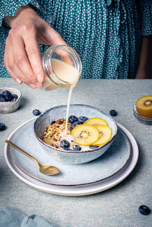 High angle of crop anonymous female adding milk into bowl with healthy muesli with blueberries and pieces of kiwi for healthy breakfast - ADSF32740