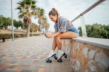 Full body side view of positive female in roller skates surfing cellphone while sitting on border on street with green trees - ADSF32742