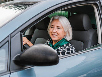 Fröhliche ältere Frau in elegantem Outfit fährt modernes glänzendes Auto in sonnigem Tag und schaut weg - ADSF32747