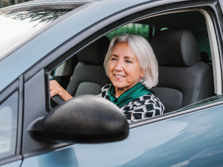 Cheerful senior female in elegant outfit driving modern shiny car in sunny day and looking away - ADSF32747