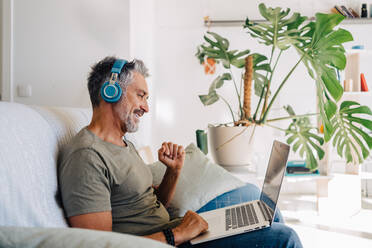 Seitenansicht eines reizenden älteren Mannes mit Kopfhörern, der mit seinem Laptop in einer modernen Loftwohnung auf dem Sofa sitzt und Musik hört - ADSF32754