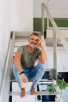 Älterer, fröhlicher, barfüßiger Mann, der auf einer Leiter sitzt und in die Kamera schaut, während er sich in einer hellen Wohnung auf die Hand stützt - ADSF32758