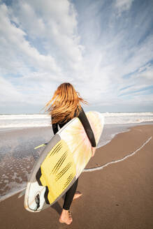 Full body back view of sportive female surfer in black wetsuit walking on wet shore near waving sea during training - ADSF32767