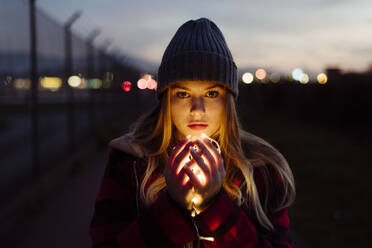 Pretty blonde young woman with wool hat holding a light garland of lights looking at the camera on the street - ADSF32768